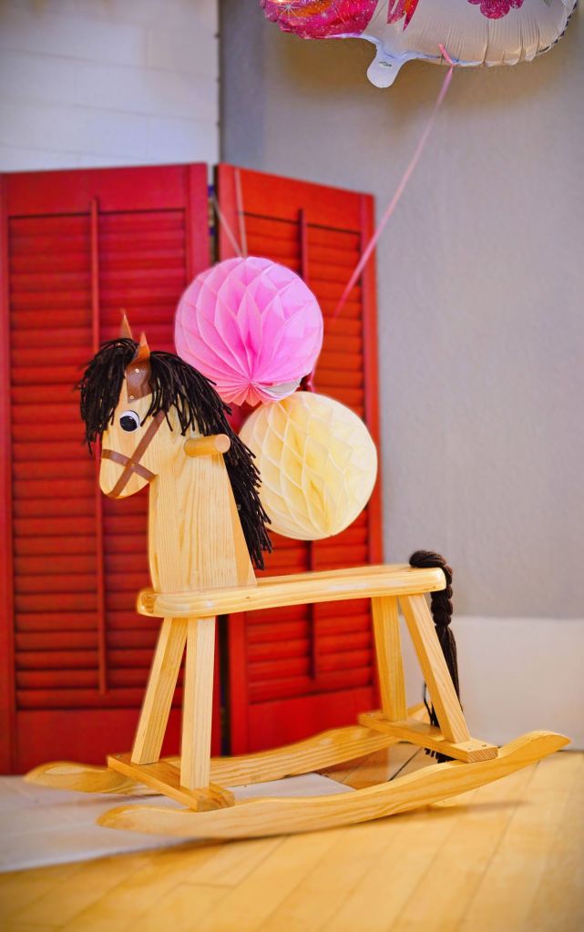 A classic light wood rocking horse with a dark yarn mane and tail, decorated with a pink honeycomb paper ball and a yellow balloon, set against a red shutter screen background.