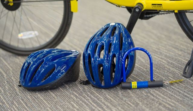 Two glossy blue bicycle helmets and a blue U-lock on a carpeted floor next to a yellow ofo bicycle.