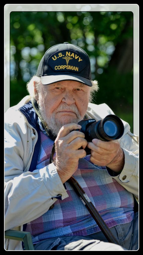 An elderly man with a grey beard, wearing a U.S. Navy Corpsman cap, holds a Nikon DSLR camera while sitting outdoors.