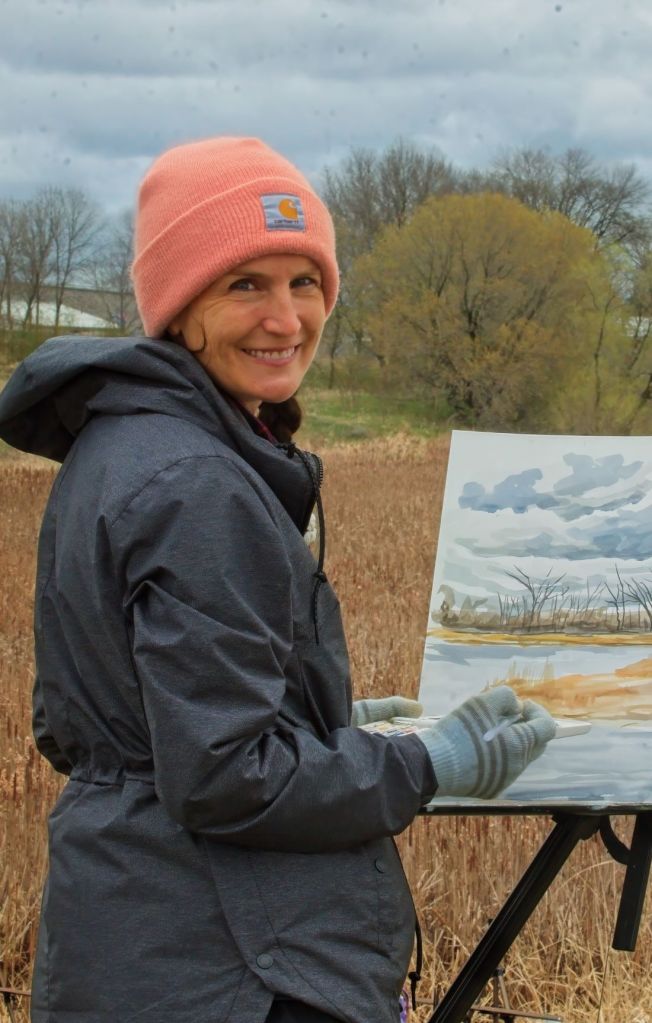 A smiling woman in a dark grey jacket and a peach-colored Carhartt beanie paints a landscape watercolor on an easel outdoors in a grassy field.