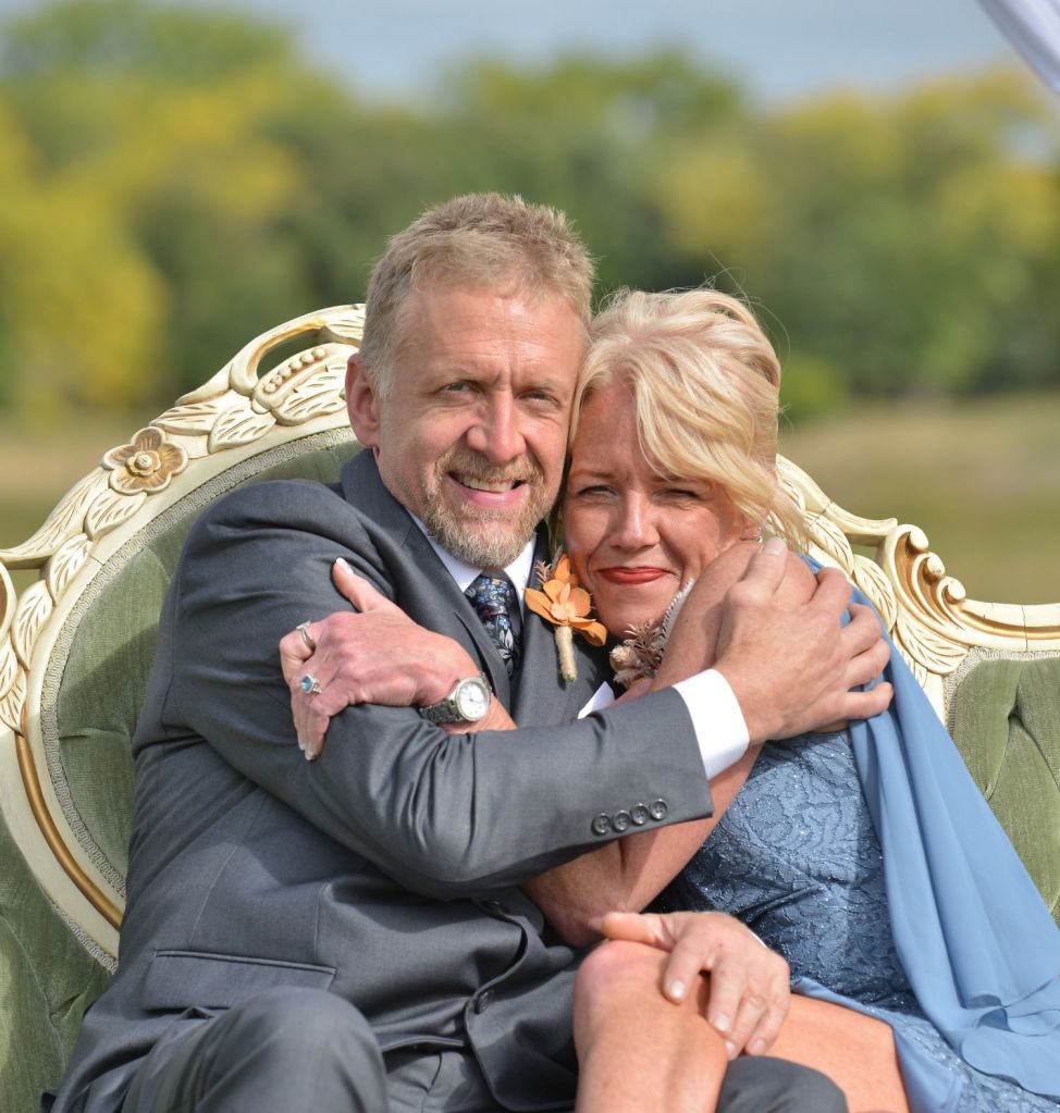 A smiling man in a grey suit and a blonde woman in a blue lace dress embracing while sitting on an ornate green and gold vintage sofa outdoors.