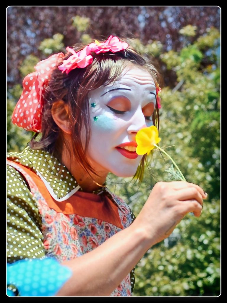 Close-up profile photograph of a smiling woman dressed as a clown with white face paint, blue cheek accents, and a red polka-dot hair bow, holding and smelling a small yellow poppy flower outdoors.