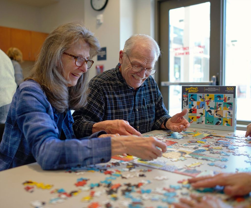 A smiling couple working together on a colorful jigsaw puzzle at the Fergus Falls Library, photographed by Lance Albers. Laughing while assembling a 500-piece "Something's Amiss!" puzzle near a bright window.