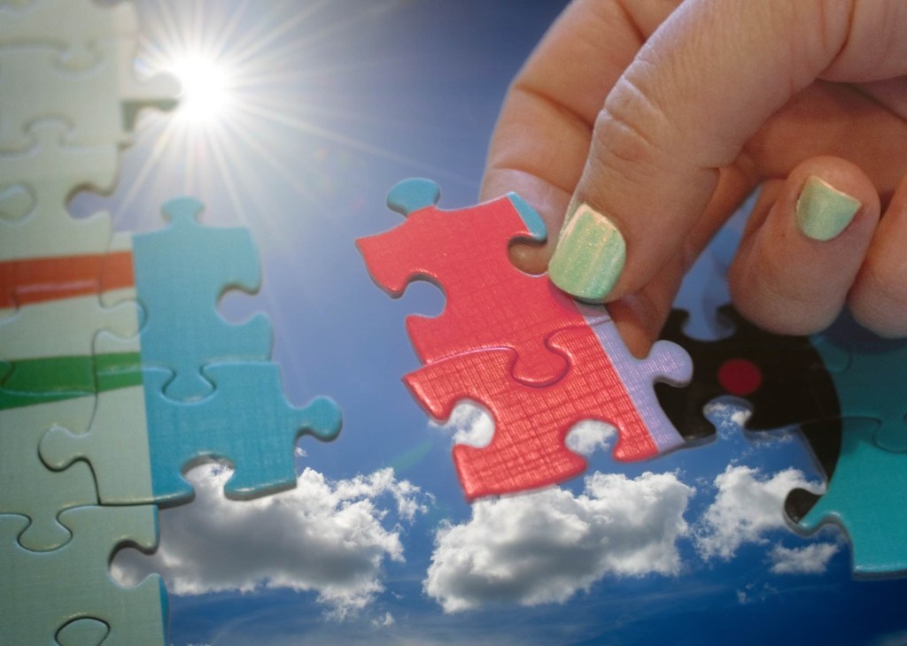 Close-up of a hand with green painted fingernails placing a red jigsaw puzzle piece into a puzzle structure, overlaid with a bright blue sky, white clouds, and a brilliant sunburst.