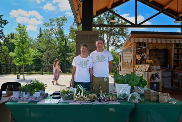 Ramona Heitmann and Doug Davidson standing behind their vibrant R&D Gardens fresh produce display at the Spies Pavilion farmers market in Fergus Falls Minnesota.