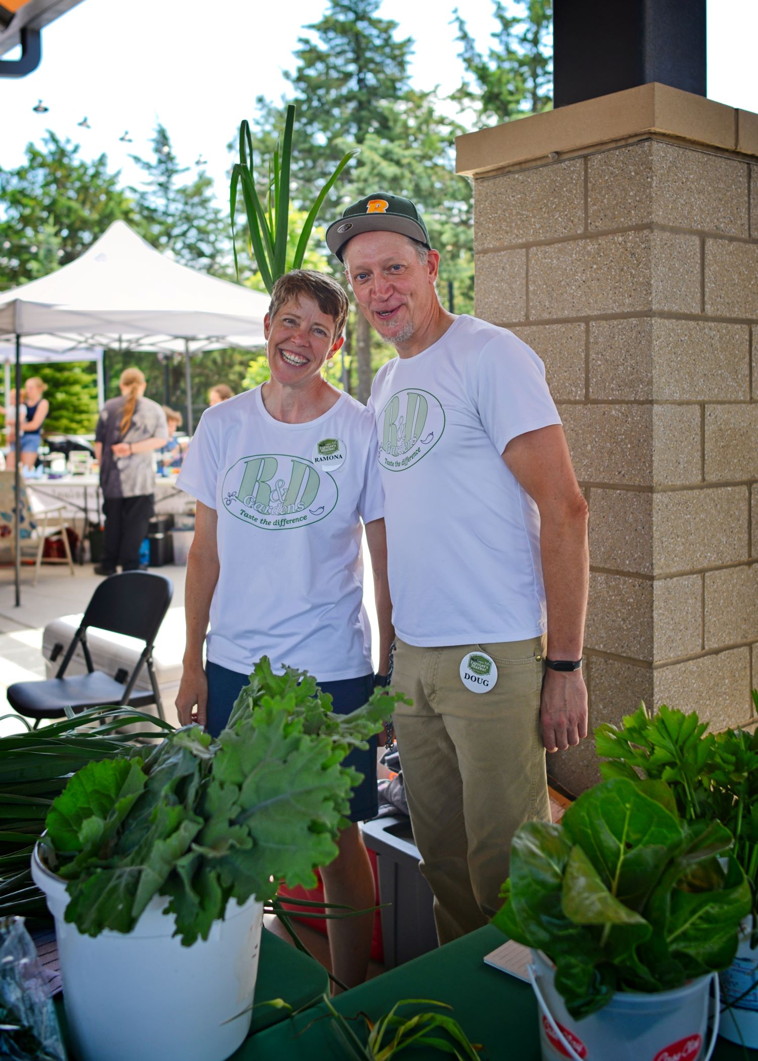 Close up portrait of smiling owners Ramona Heitmann and Doug Davidson wearing matching white R&D Gardens shirts behind their produce table at the Fergus Falls Farmers Market Minnesota.