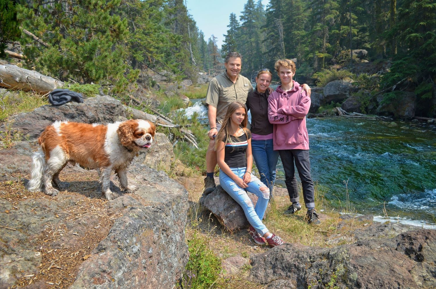 A family of four and their Cavalier King Charles Spaniel posing on rocky terrain next to a rushing forest river.