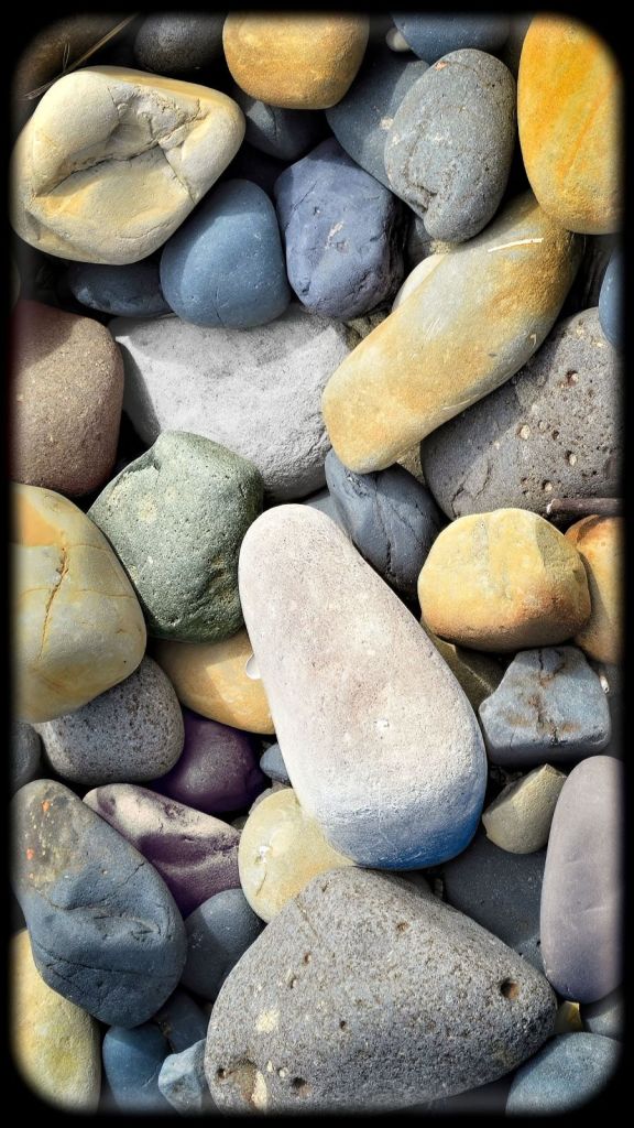 A top-down abstract view of smooth, rounded river stones and pebbles in various shades of blue, gray, cream, yellow, and purple.