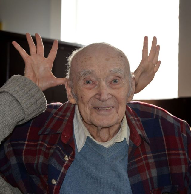 Portrait of Roger Thompson celebrating his 100th birthday as someone playfully makes deer or moose antlers behind his head, photographed by Lance Albers.