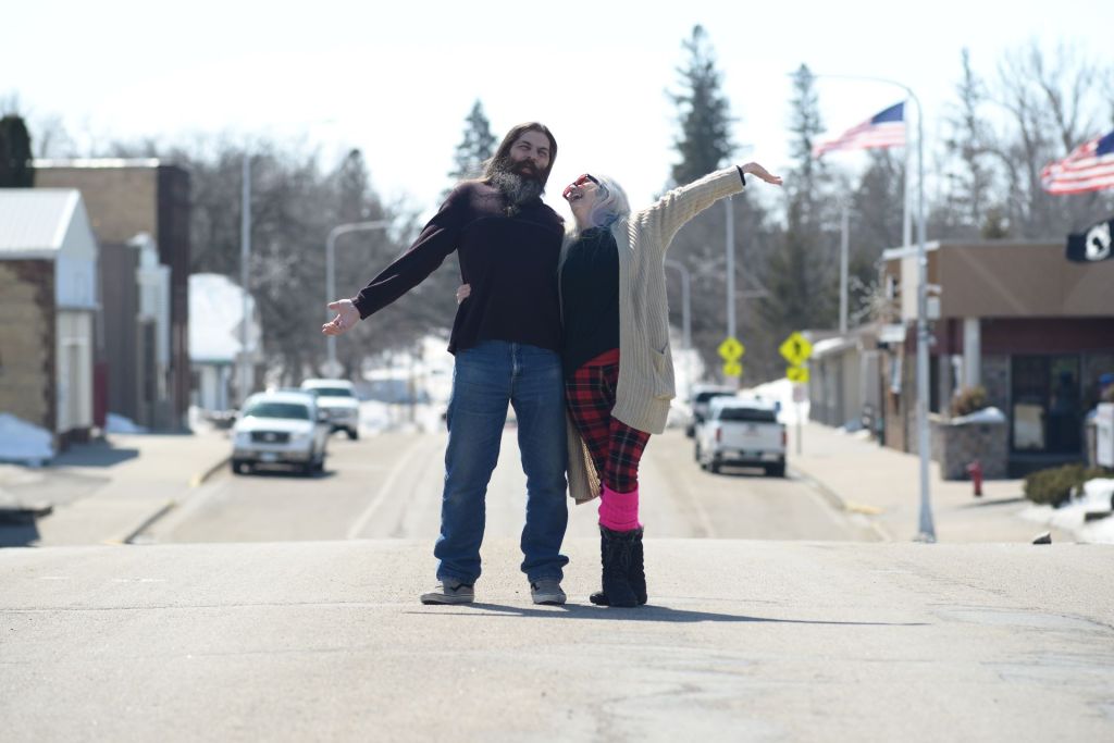 An emotional outdoor engagement portrait of a couple celebrating a milestone of safety and healing by standing joyfully with outstretched arms in the center of an empty small-town street, photographed by Lance Albers.