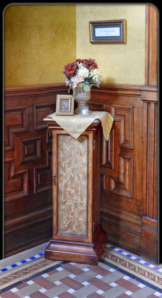 A decorative wooden pedestal cabinet holding a floral arrangement and a framed picture, positioned in a corner with historic wood wainscoting and tiled flooring.