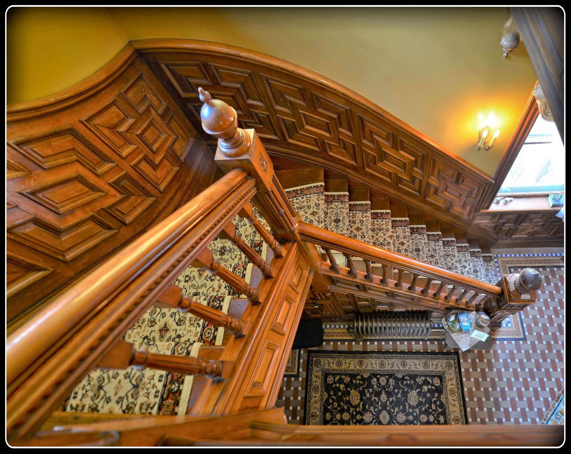 High angle view of a historic carved wood grand staircase with geometric wall paneling and a carpet runner inside the Samuel Dearing House in St Paul.