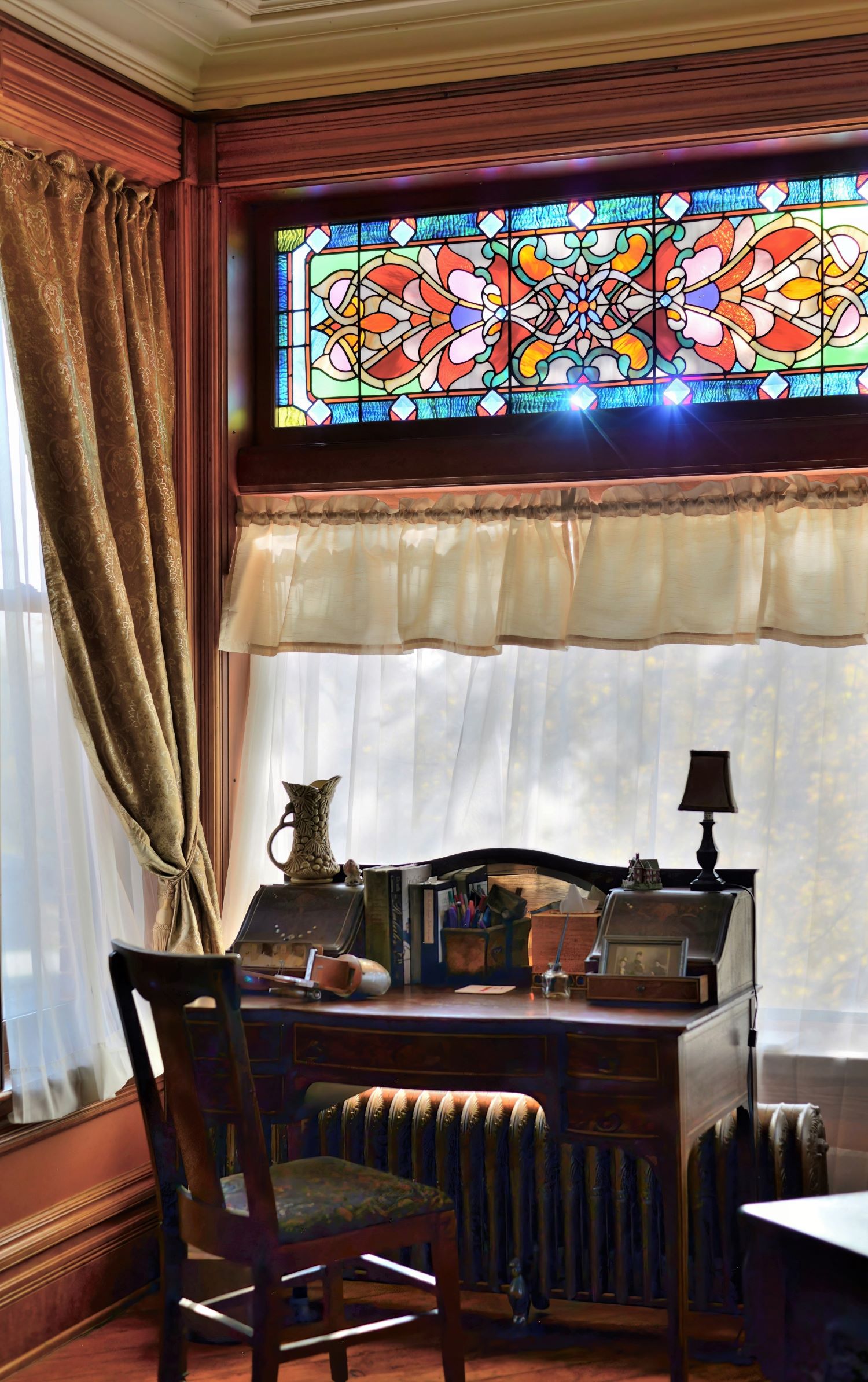 Antique wooden writing desk and matching chair set against a window with a large, colorful stained-glass transom overhead, framed by elegant gold curtains in a historic interior.