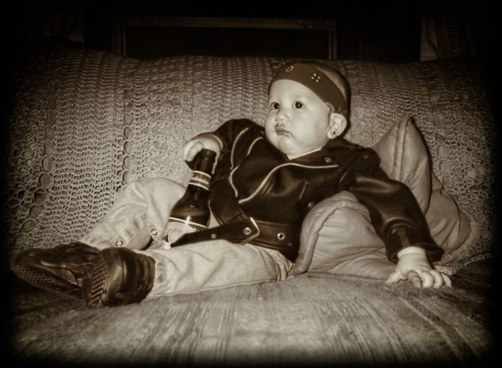 Sepia-toned portrait of a relaxed baby dressed as a biker for Halloween, 2004. He is wearing a black leather jacket, jeans, boots, and a bandana, leaning against cushions while sticking his finger down a glass Budweiser beer bottle.