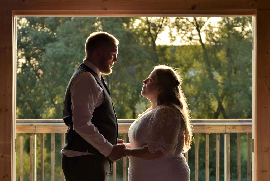 A man in a vest and a woman in a white lace dress hold hands and look at each other, framed by a wooden doorway with a backlit, natural green background.