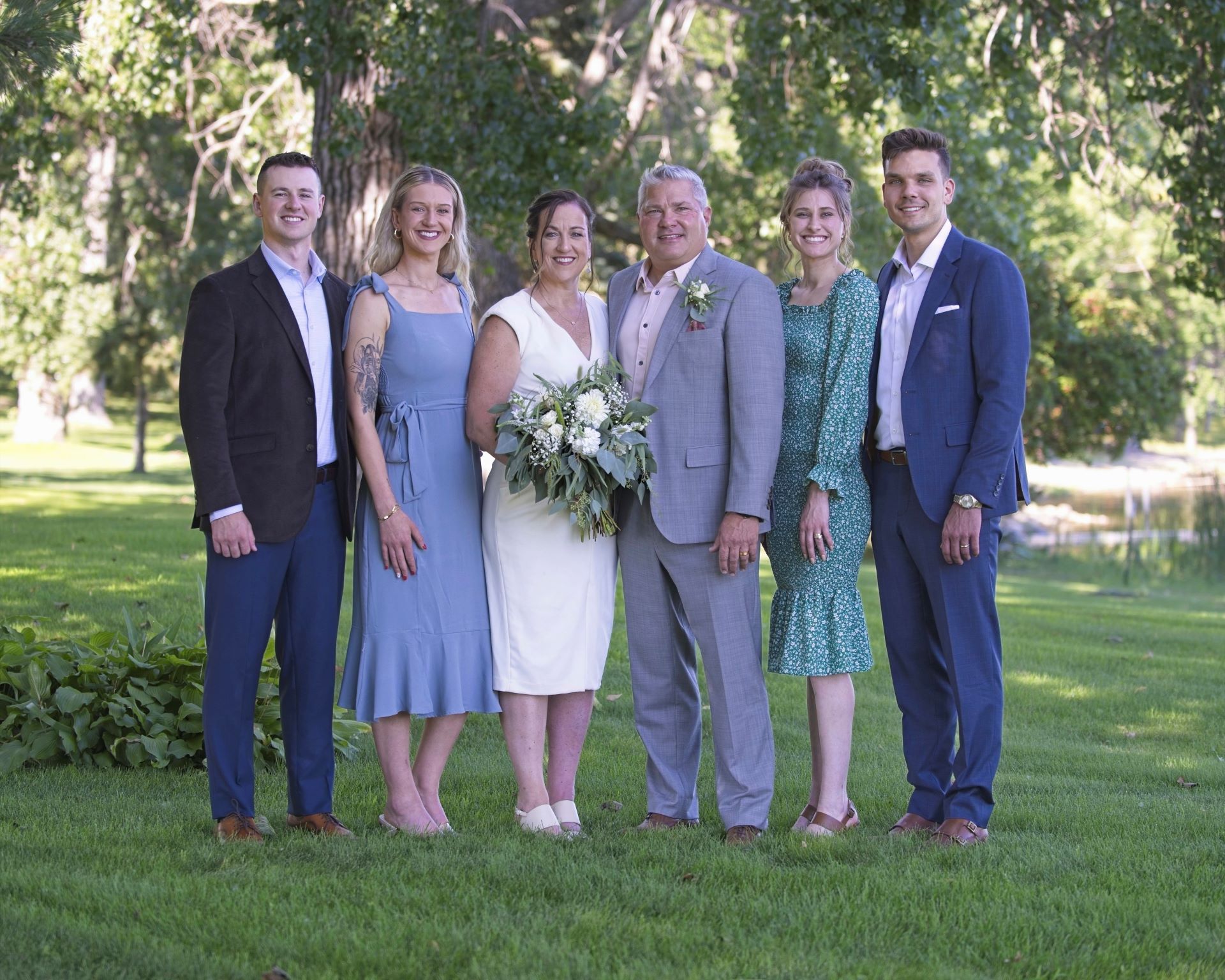 An outdoor family wedding portrait featuring a bride and groom with their family on a green lawn by a lake in Otter Tail County, Minnesota.