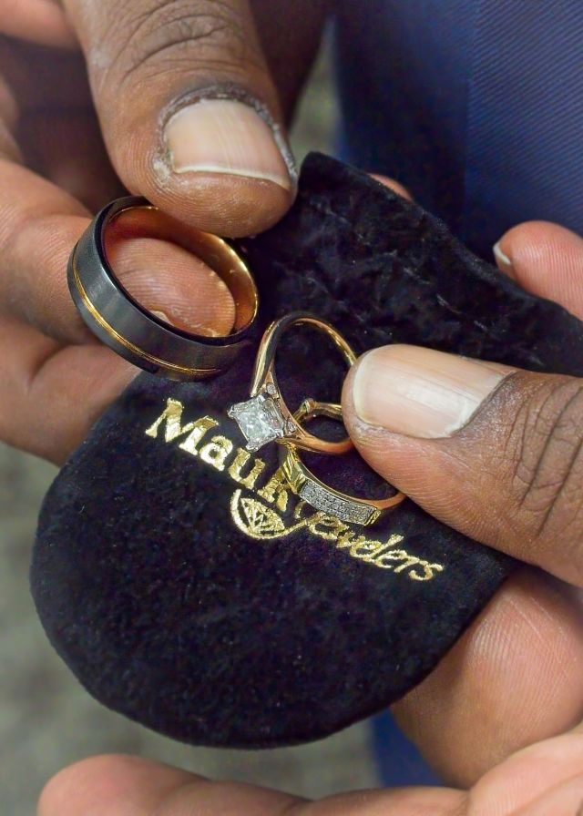 Close-up shot of a person holding a black velvet jewelry pouch from Mauk Jewelers displaying a sparkling princess-cut diamond engagement ring and a modern black and gold men's wedding band.