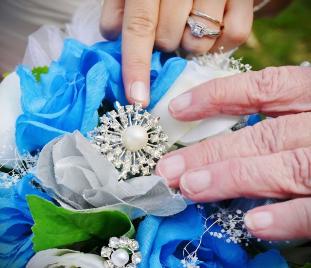 Close-up of two hands gently touching a silver starburst brooch with a central cream pearl accent nestled inside a bridal bouquet of bright blue, white, and silver artificial roses.