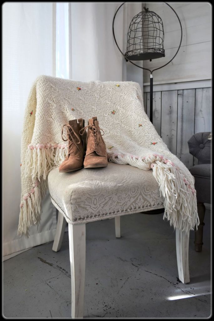 A pair of brown suede lace-up heeled ankle boots rests on an upholstered white accent chair draped with an embroidered cream knit blanket with long fringe.