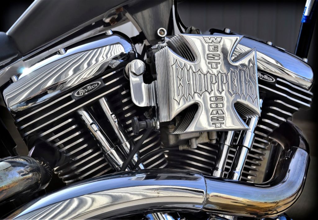 Macro photograph of a polished chrome motorcycle engine with RevTech branding, focusing on an engraved, cross-shaped West Coast Choppers air intake assembly.