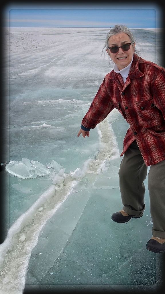 A woman in a red plaid jacket points nervously at a large pressure ridge crack in the thick blue ice of Lake of the Woods, MN during winter.