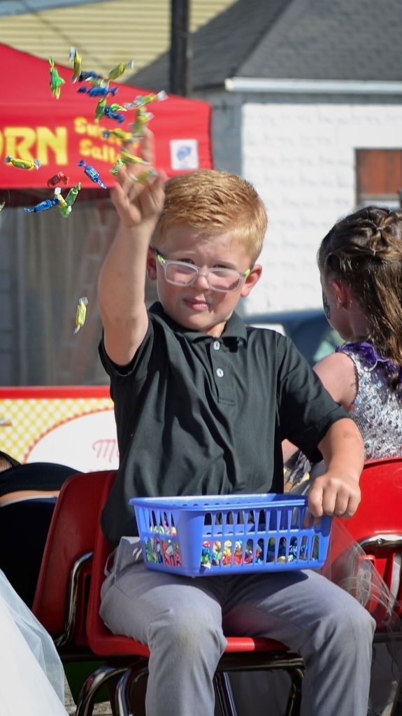 A young boy wearing glasses and a black polo shirt is throwing a handful of colorful wrapped candies from a blue plastic basket on his lap during a parade.