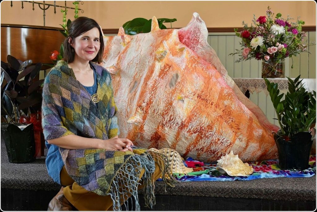 A woman sitting on a stage next to a massive, colorful papier-mache conch shell sculpture and an array of real seashells and floral arrangements.