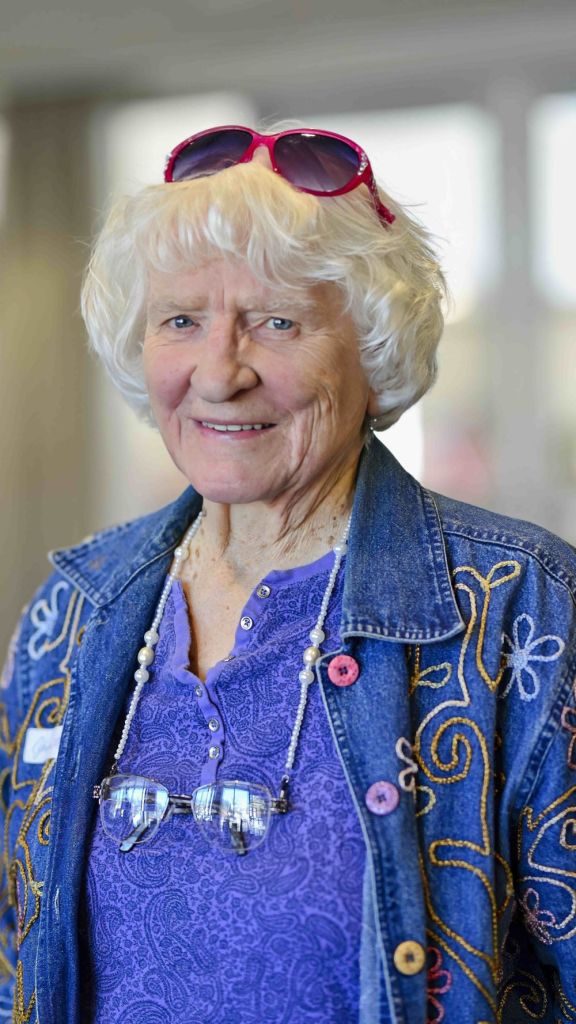 A smiling senior woman with white hair wears a decorated denim jacket and pink sunglasses at the Fergus Falls Library.