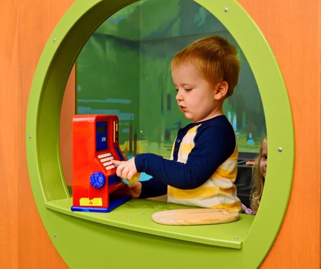 A young boy presses buttons on a toy cash register inside a circular play window at the library.