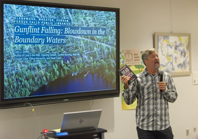 Minnesota author Cary J. Griffith speaking into a microphone at the Fergus Falls Public Library while holding his mystery novel "Dead Catch" next to a large digital screen displaying "Gunflint Falling: Blowdown in the Boundary Waters."