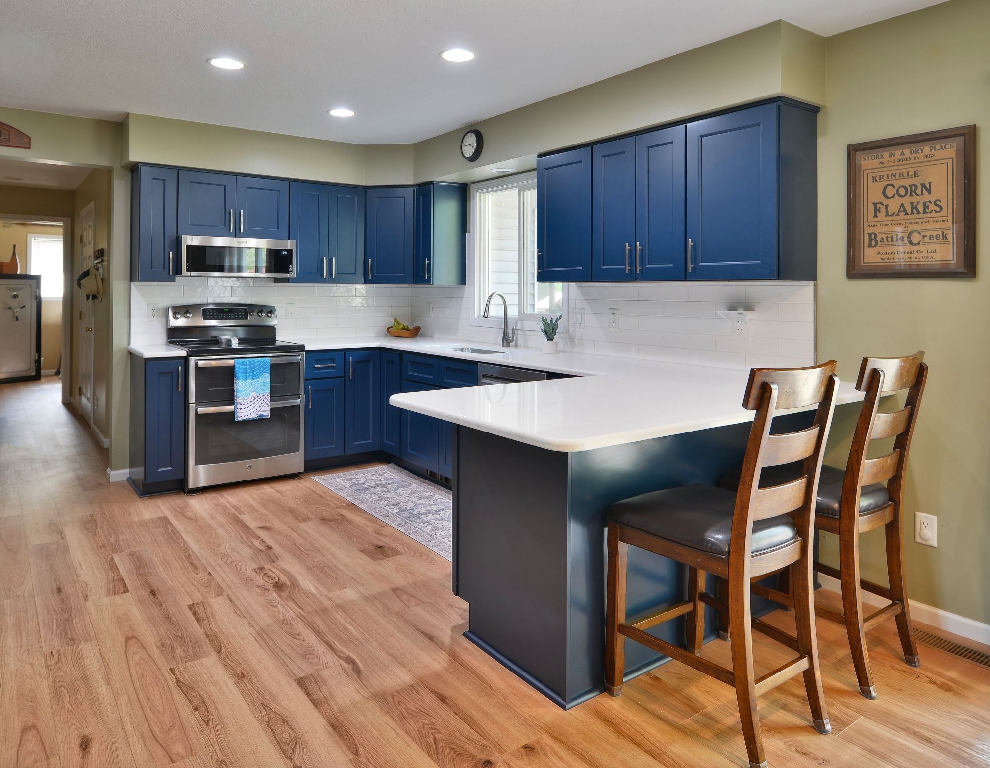 Professional interior photograph of a spacious, remodeled kitchen featuring deep navy blue cabinets, white quartz countertops, stainless steel appliances, and warm hardwood floors.