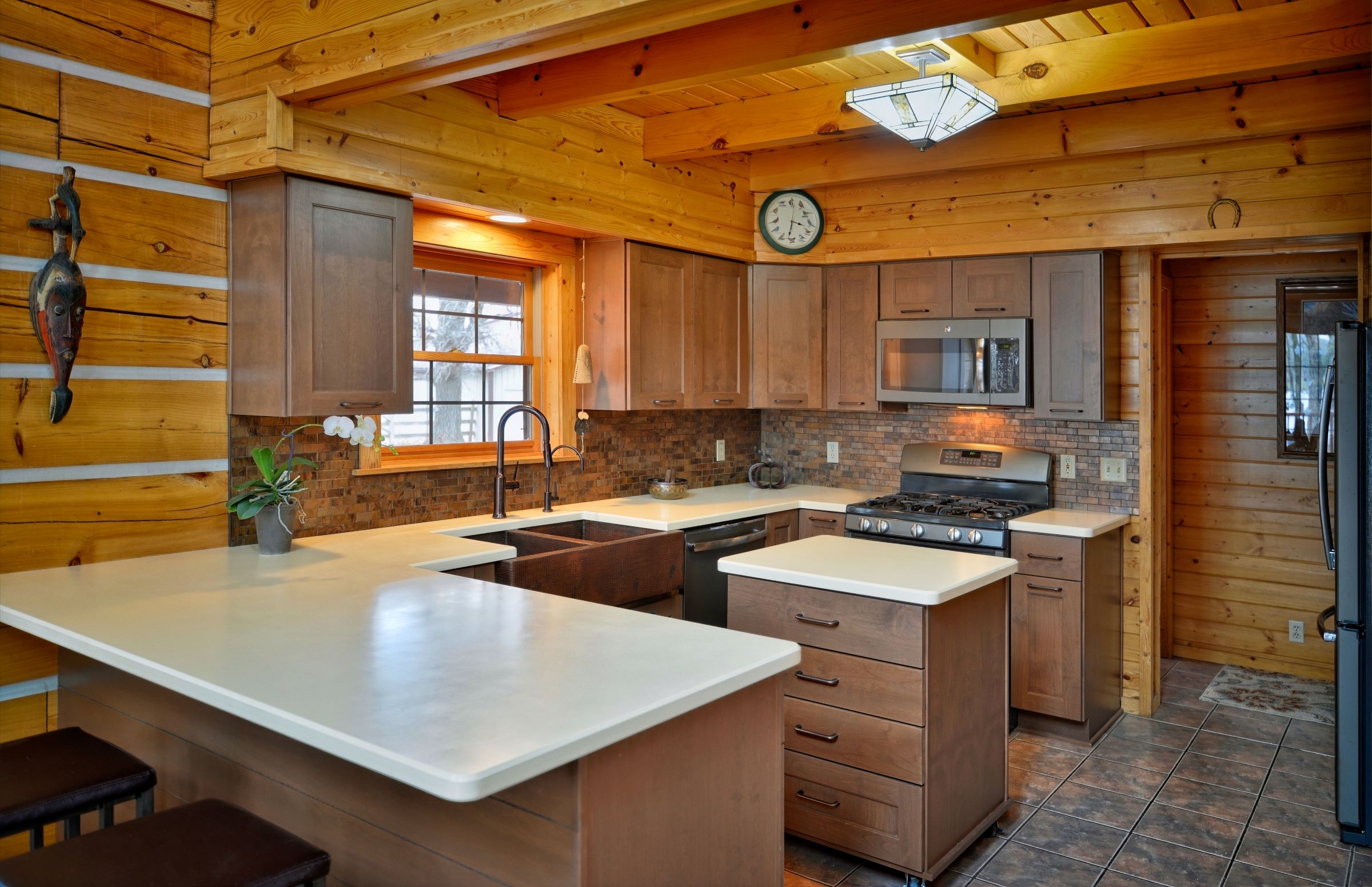 Professional interior photograph of a rustic log home kitchen featuring custom muted brown cabinets, white solid-surface countertops, an island, and exposed log walls.