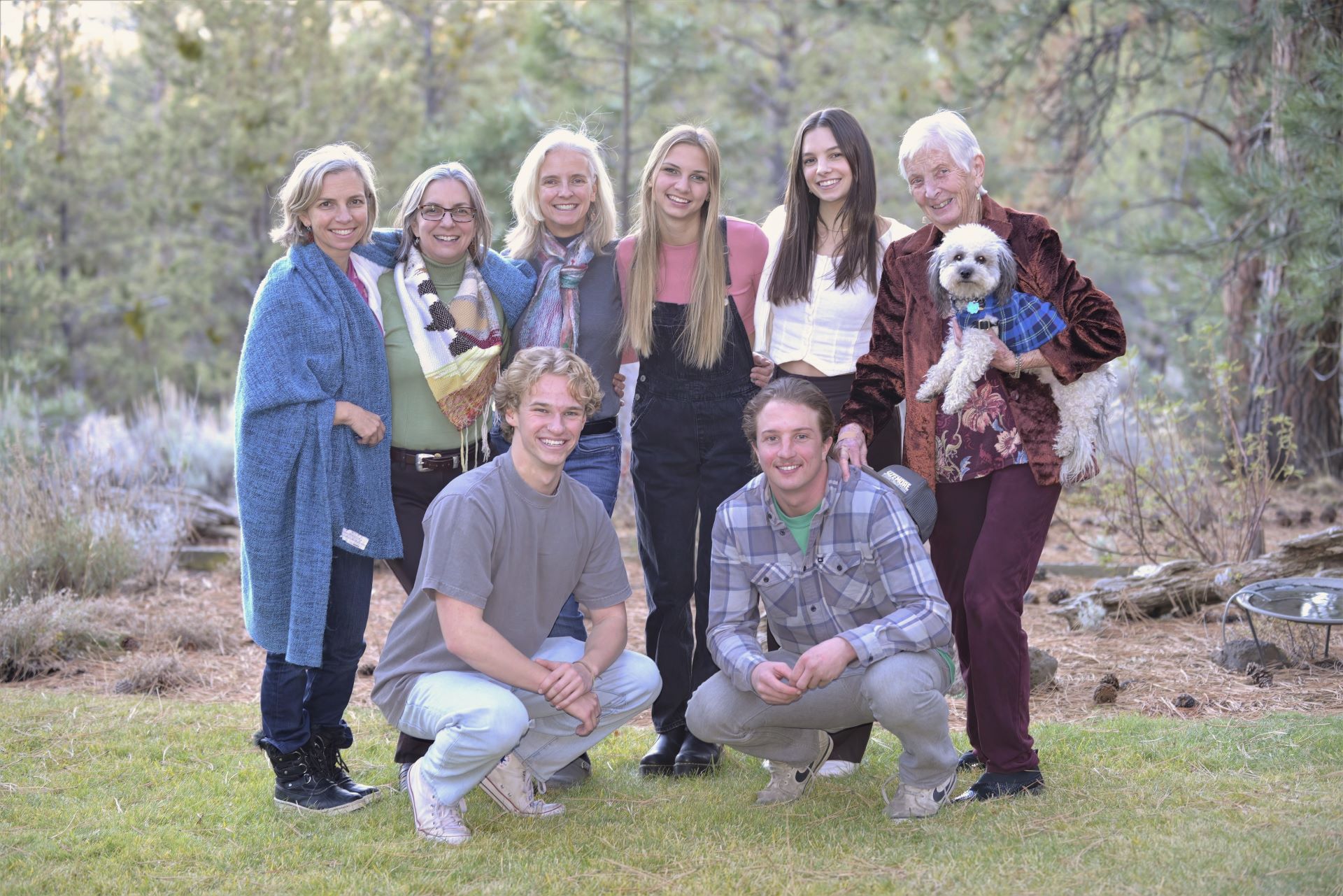 An outdoor multi-generational family portrait of eight people and a small dog standing and crouching on a green lawn surrounded by soft pine trees.