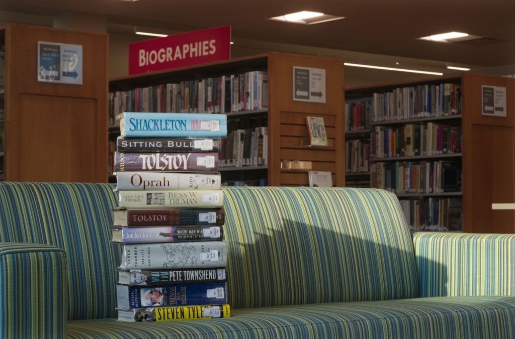 Stack of biography books including Shackleton and Tolstoy on a green and blue striped couch inside a public library.