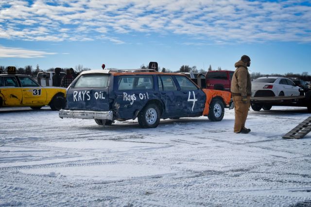 • A driver stands on the snow-covered ice of Bass Lake next to a custom number 4 Chevrolet Malibu station wagon prepped for ice racing.
