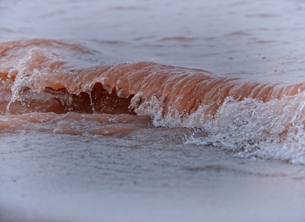 A close-up photograph of a curling, textured wave on Lake Superior tinged a deep reddish-brown color from suspended red clay sediment along the northern Wisconsin shore.