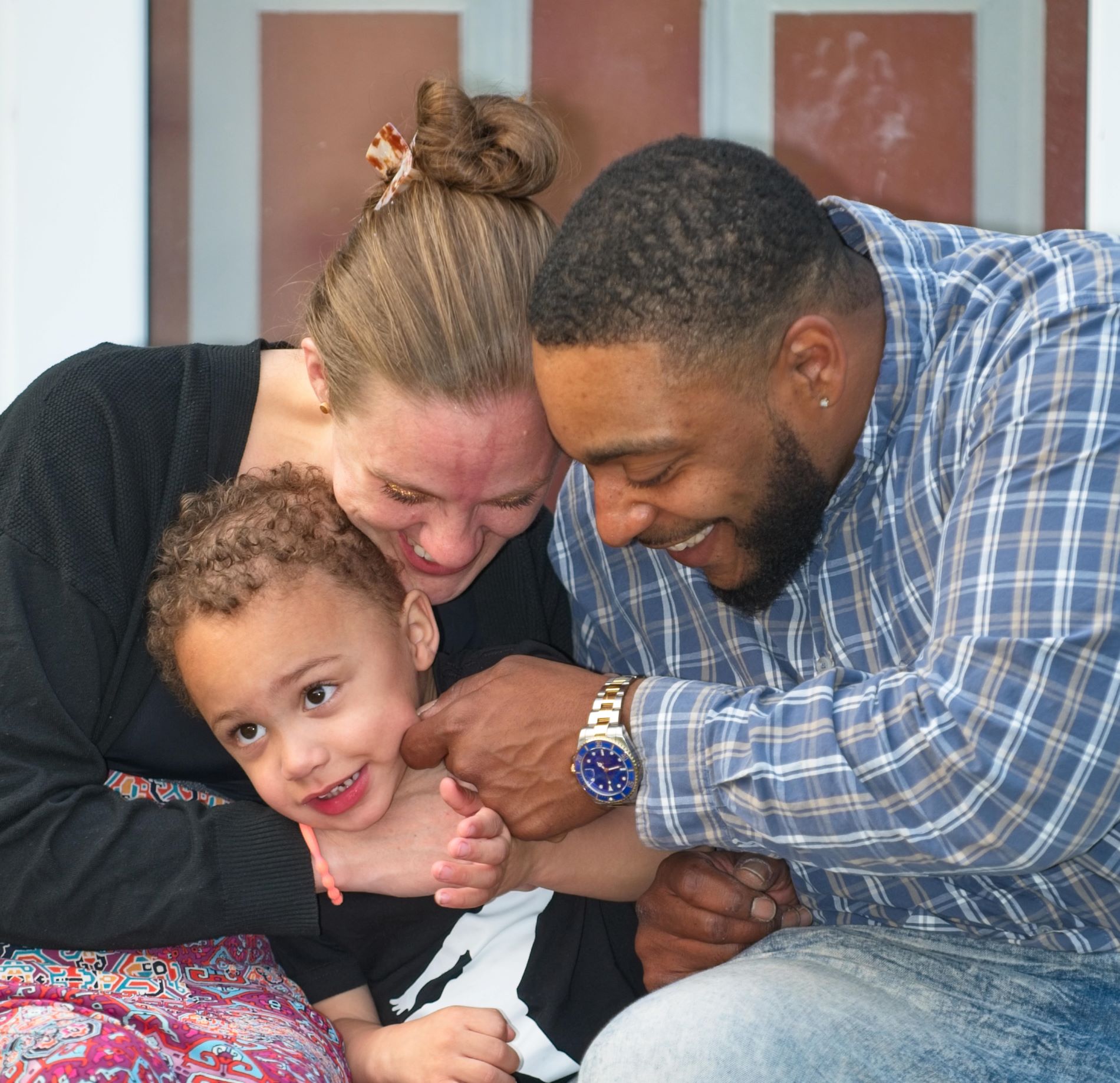 Candid close-up portrait of a laughing mother and father leaning in to embrace their smiling young child.