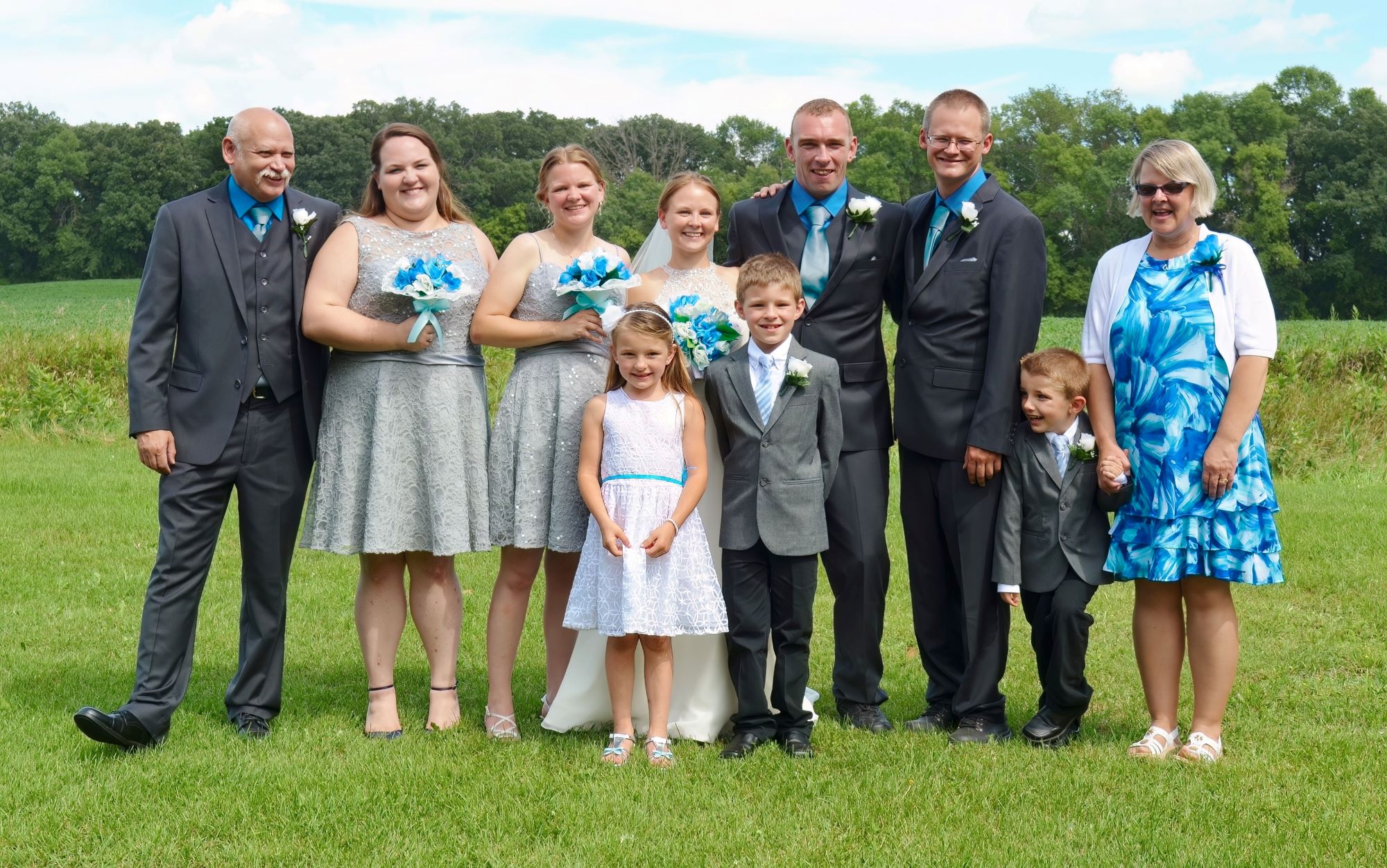 Professional outdoor wedding group photograph of a bride, groom, bridesmaids, groomsmen, and family members posing together on a green grass field with a lush treelined backdrop.