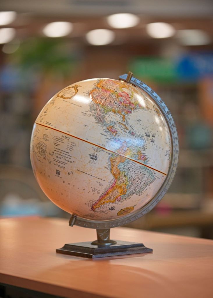 An antique-style 12-inch Replogle desktop world globe on a tiered dark base, sitting on a wooden surface inside the Fergus Falls Library with a blurred background.