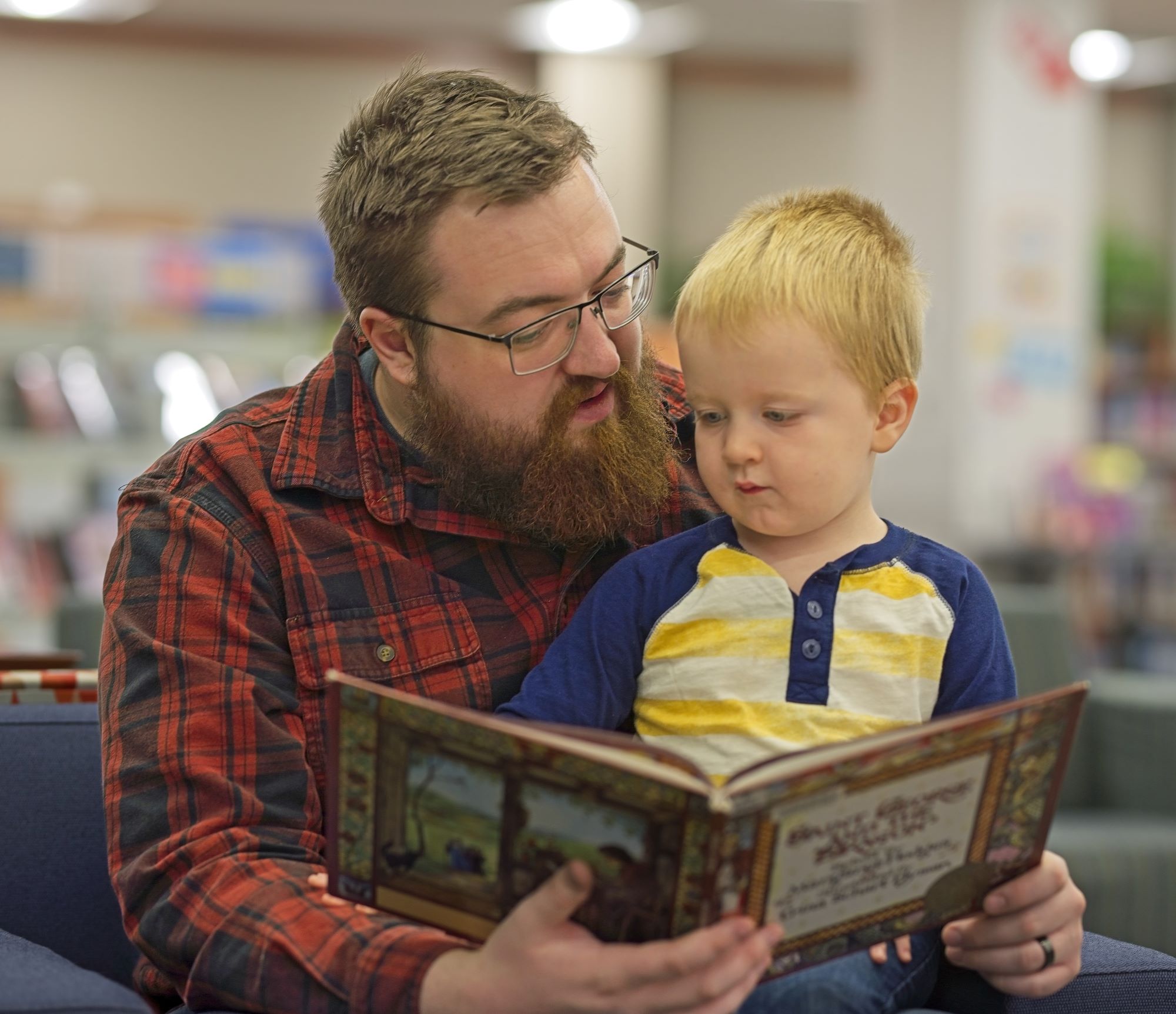 A father with a beard and glasses reads a picture book to his young son at the Fergus Falls Public Library.