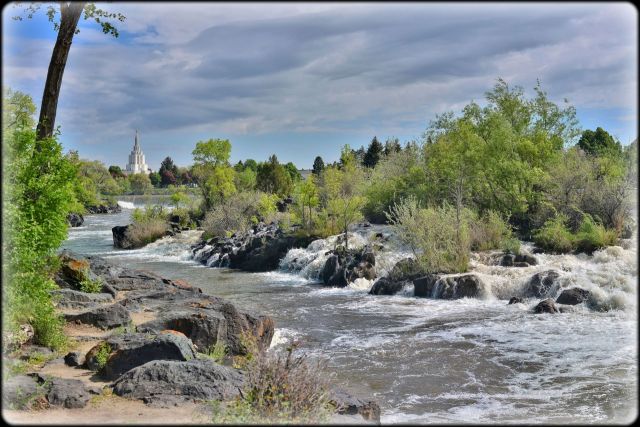 Landscape photograph of the rushing Snake River flowing over dark basalt lava rocks along the Idaho Falls Greenbelt, with the white spire of the Idaho Falls Idaho Temple visible in the background under a cloudy sky.