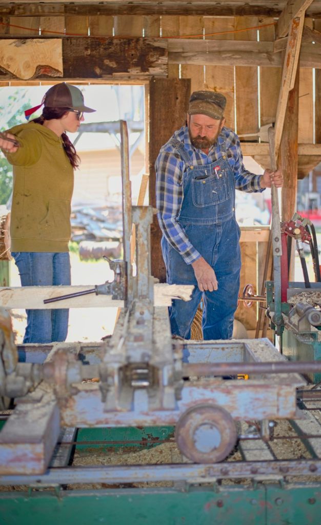 Jason Olson of Rothsay and a young assistant operating a vintage sawmill inside a rustic wooden barn, cutting fresh cottonwood lumber powered by an external steam engine at the Dalton Threshing Show.