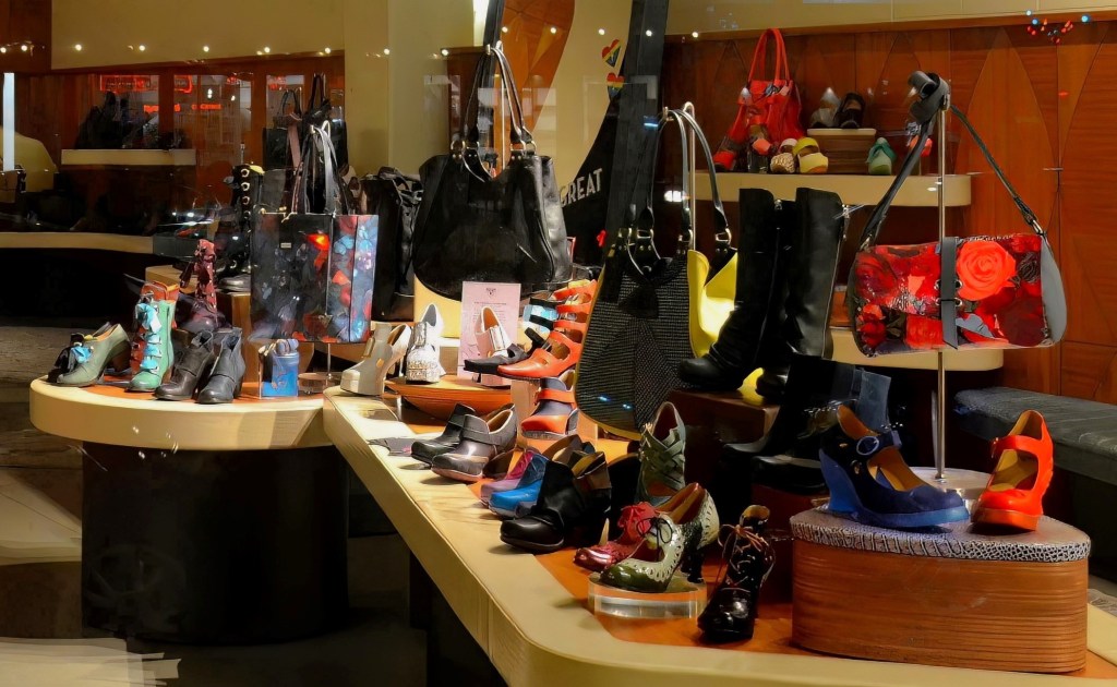 A vibrant retail display inside the John Fluevog Shoes store in the Pearl District of Portland, Oregon, showcasing a colorful collection of artistic leather boots, patterned heels, unique sandals, and floral handbags arranged on tiered wooden tables.