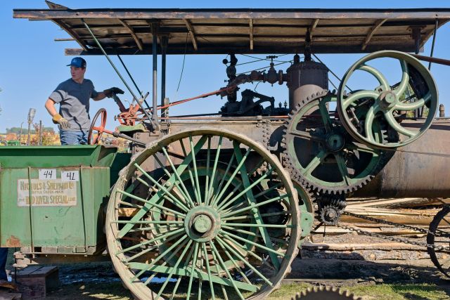 A young man operating a large, vintage green and brown Nichols and Shepard steam traction engine belted to power a sawmill at the 2026 Dalton Threshing Show.