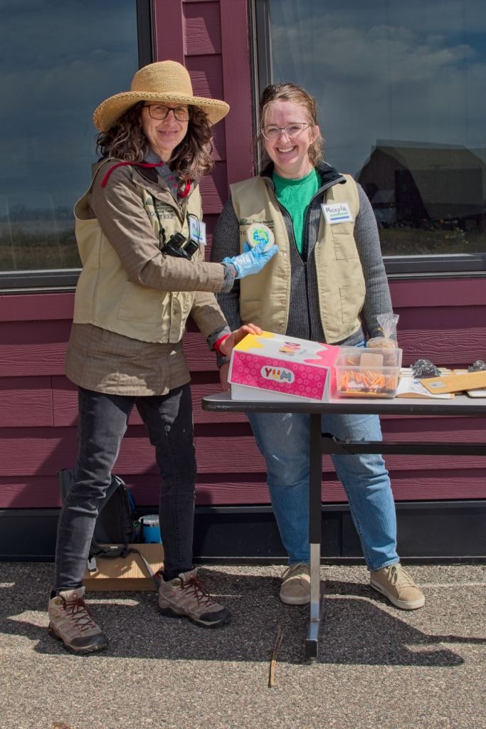 Two women smiling outside at the Prairie Wetlands Learning Center, wearing tan volunteer vests while holding up a colorful Earth Day badge next to a display table.
