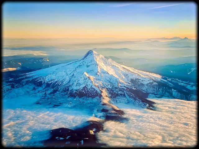 High-altitude aerial photograph of Mount Hood's snow-covered peak rising majestically above a vast, thick blanket of white clouds under a clear blue sky, as seen from an airplane window.