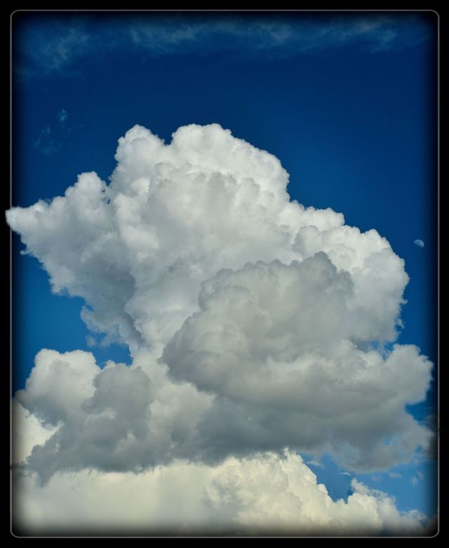 Fine art photograph of a massive, towering white cumulus cloud formation rising against a deep blue sky with a faint crescent moon visible on the right edge.