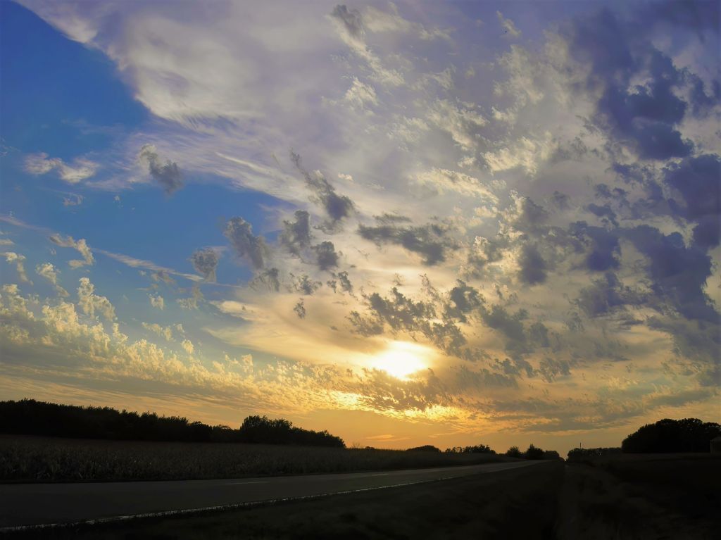 Dramatic morning sky over a quiet country road near Dalton, Minnesota, with golden sunlight breaking through scattered, textured clouds.