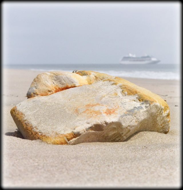 Artistic close-up of a weathered, sandy boulder with warm golden undertones resting on a sandy beach, with a blurred cruise ship on the distant misty horizon.