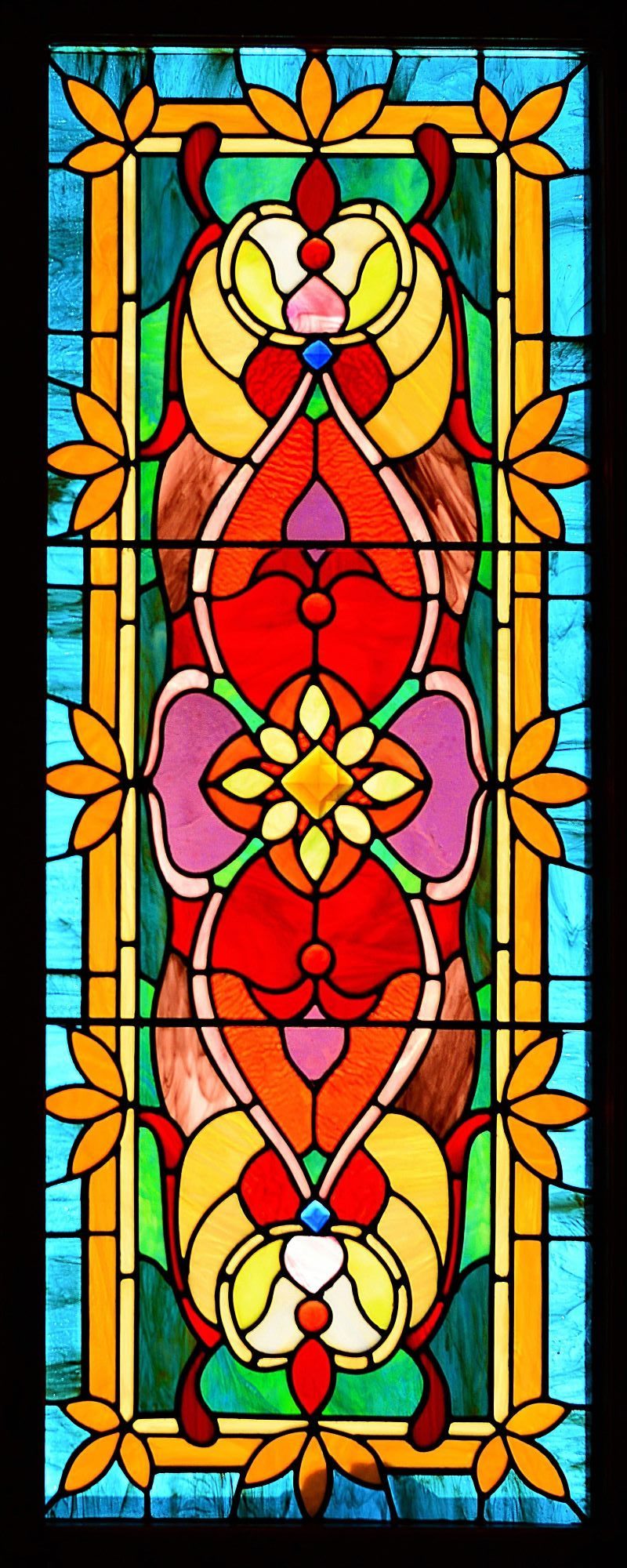 Vertical photograph of a symmetrical, multicolored Art Nouveau stained glass window panel with vibrant red, yellow, green, and blue glass segments.